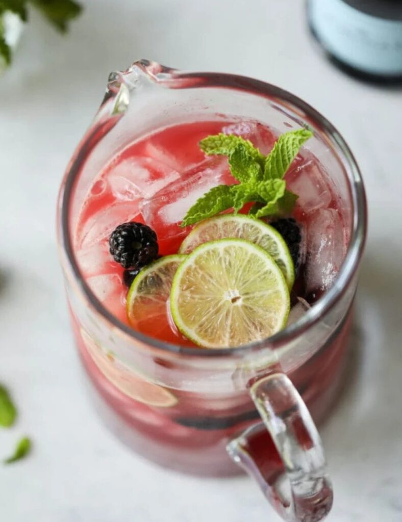 a glass pitcher of red drink with limes and mint