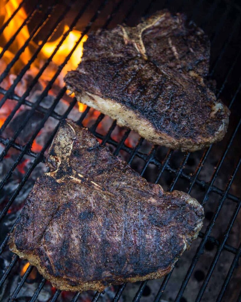 Two steaks sizzling on a grill, surrounded by vibrant flames.