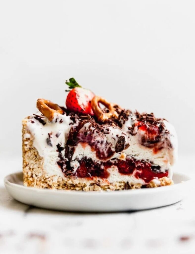 A slice of strawberry ice cream cake topped with chocolate and crunchy pretzel pieces.

