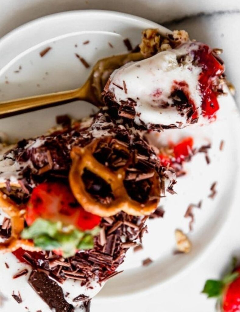 A slice of strawberry ice cream pie topped with pretzels and drizzled with chocolate sauce on a white plate.
