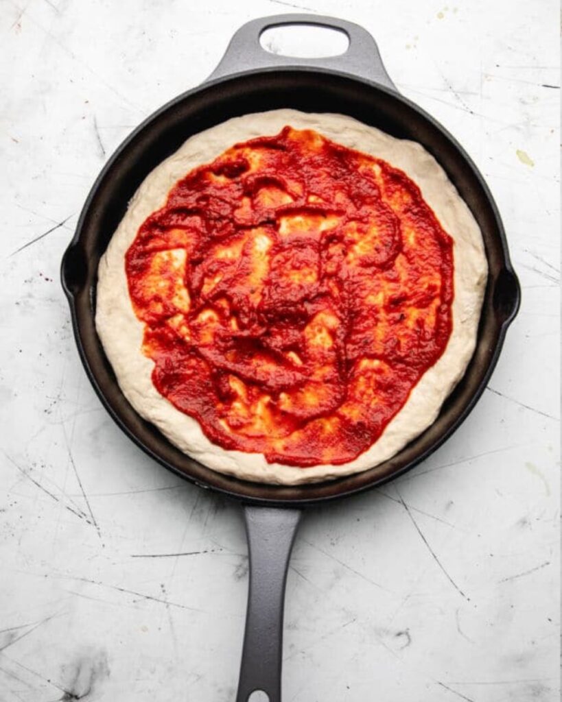 A pizza in a cast iron skillet rests on a marble counter, showcasing its golden crust and melted cheese.