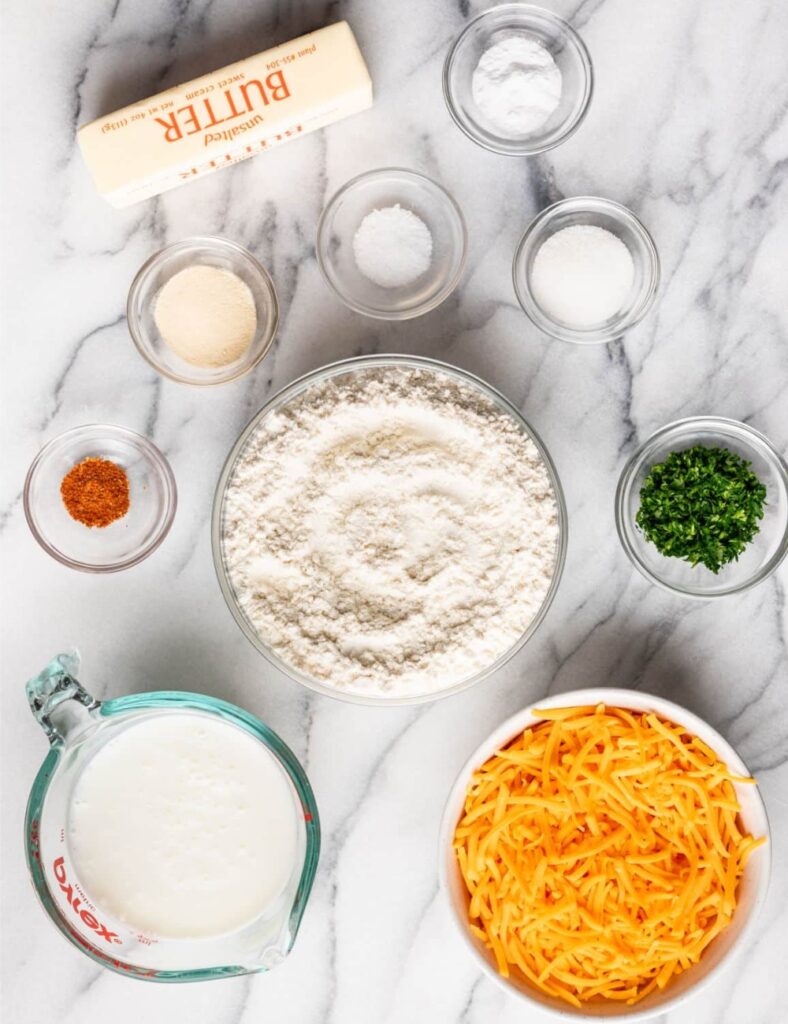 2 cups all-purpose flour, 
1 tablespoon baking powder, 
1 teaspoon garlic powder, 
1/2 teaspoon salt, 
1/4 teaspoon cayenne pepper, 
1/2 cup unsalted butter, cold and cubed, 
1 cup shredded cheddar cheese, 
3/4 cup milk, 
1 tablespoon fresh parsley, chopped, 
2 tablespoons melted butter, 
1/2 teaspoon garlic powder (for topping)
