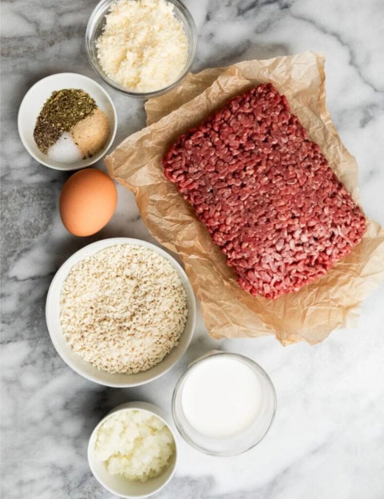 Ingredients for meatballs arranged on a marble counter, including ground meat, herbs, and spices.

