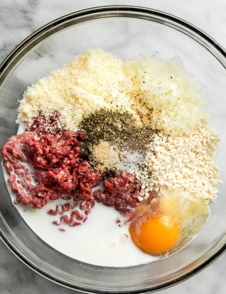 Ingredients for meatballs arranged in a bowl, including ground meat, breadcrumbs, herbs, and spices.