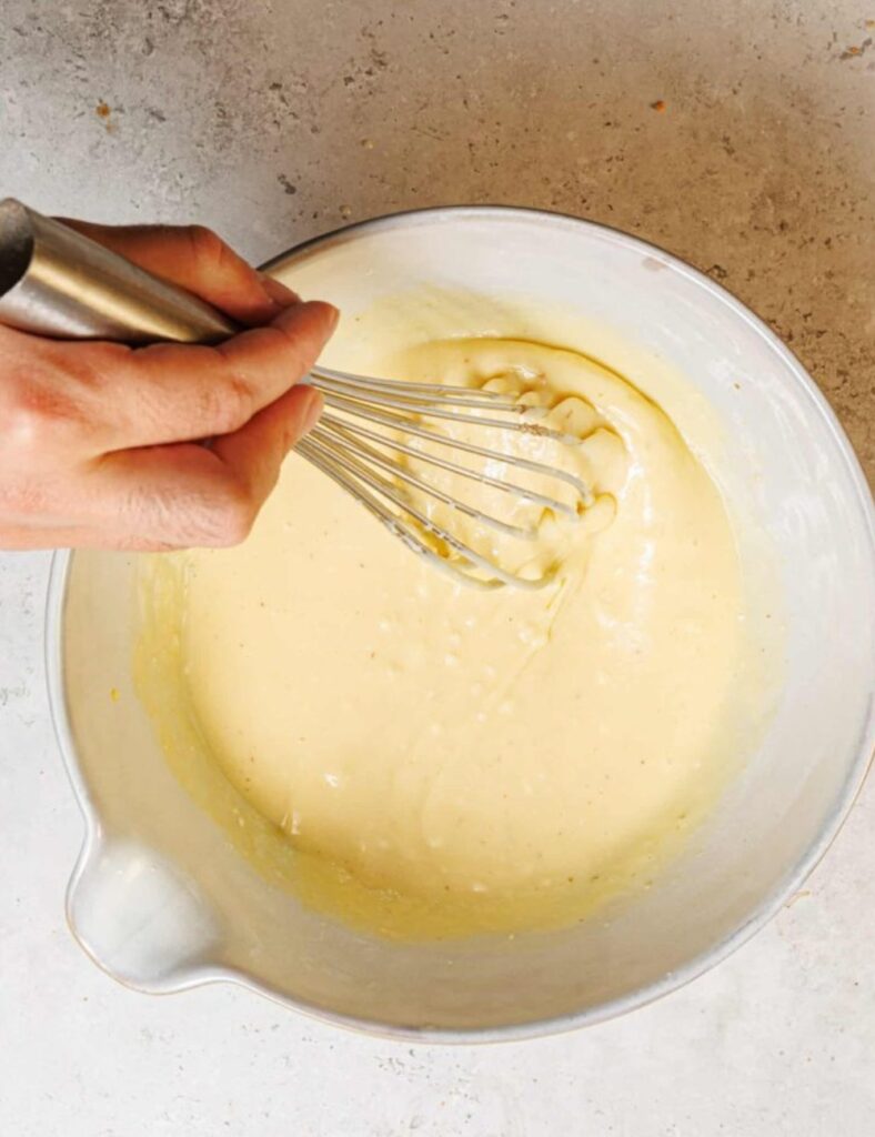 A hand using a whisk to mix batter in a bowl, with a focus on the motion and texture of the mixture.