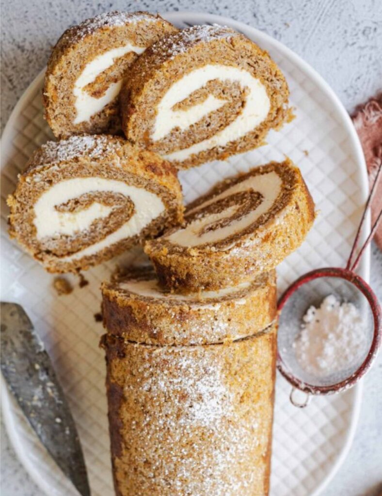 Pumpkin roll cake filled with cream cheese frosting, elegantly swirled and dusted with powdered sugar on a white plate.


