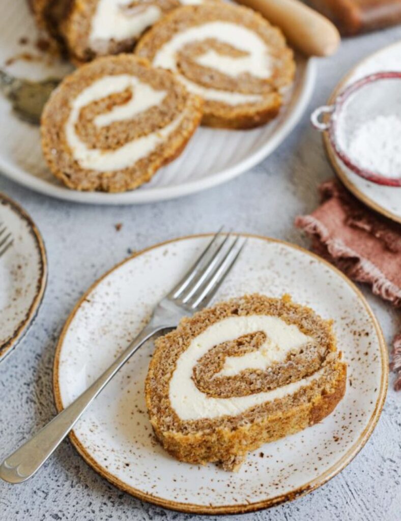 Pumpkin roll cake filled with cream cheese frosting, elegantly swirled and dusted with powdered sugar on a white plate.

