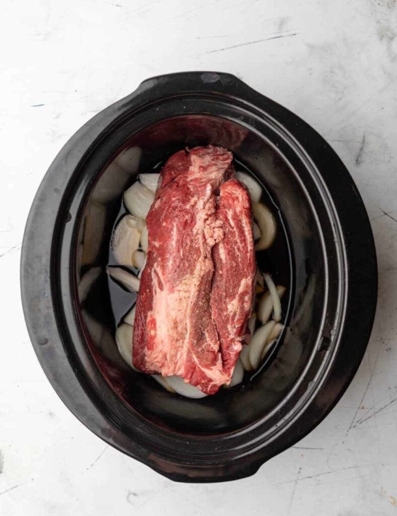 A piece of meat cooking in a slow cooker, surrounded by vegetables and broth.s

