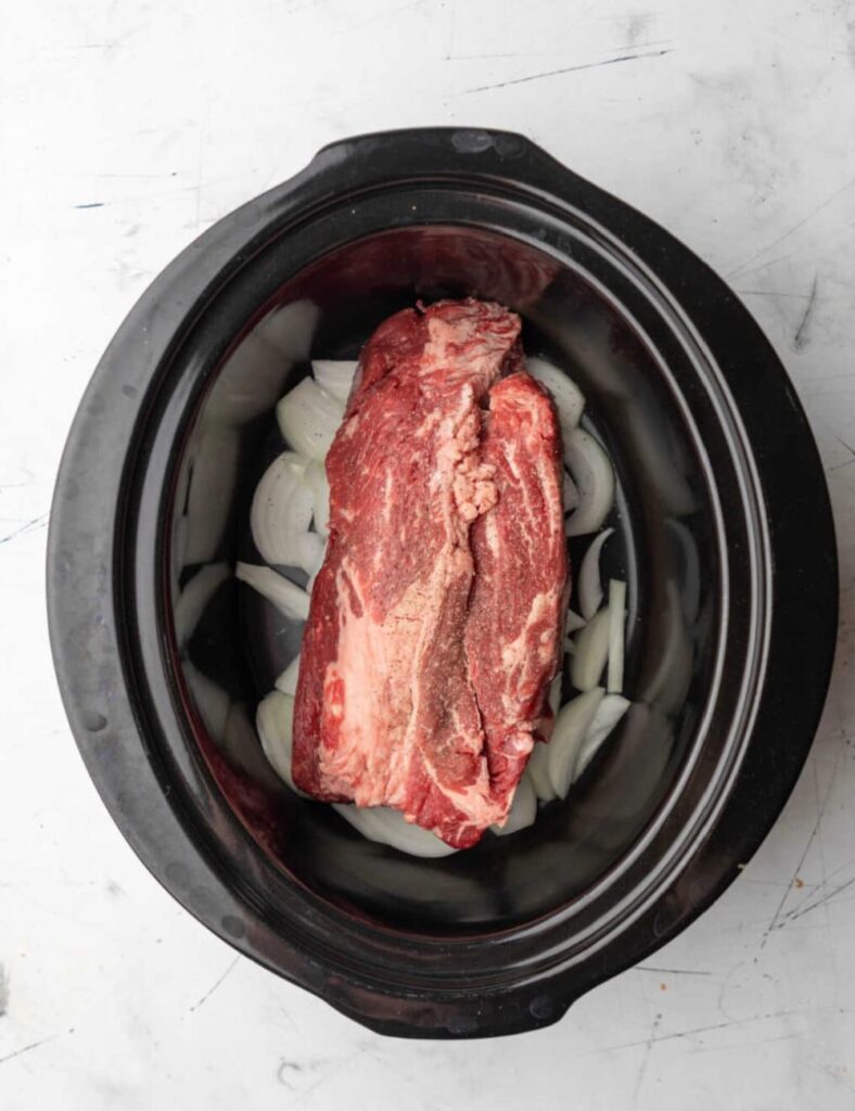 A piece of meat cooking in a slow cooker, surrounded by vegetables and broth.
